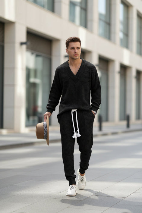 A man modeling a black cable-knit lounge set consisting of a V-neck long-sleeve sweater and matching jogger pants with a white drawstring, 