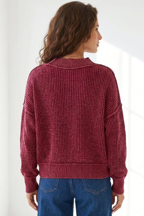 A woman viewed from the back wearing a ribbed knit sweater in a deep burgundy red, paired with blue jeans, against a bright white background.