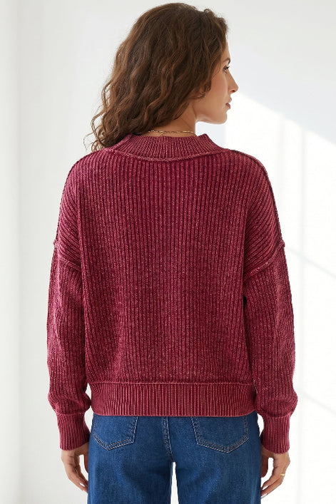 A woman viewed from the back wearing a ribbed knit sweater in a deep burgundy red, paired with blue jeans, against a bright white background.