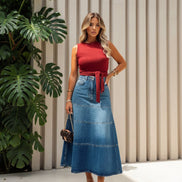 A woman stands against a white textured wall wearing a sleeveless red mock-neck top with a tie waist and a long blue denim midi skirt. 