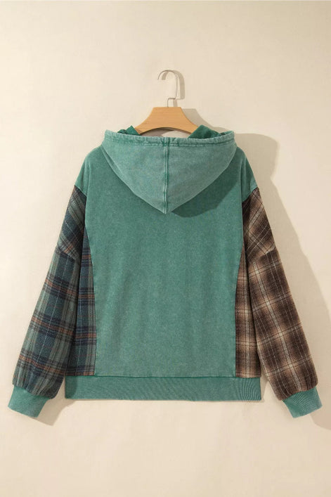 A teal green hooded sweatshirt hanging on a wooden hanger against a plain beige wall, featuring plaid patterned sleeves.