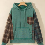 A teal green hooded sweatshirt hanging on a wooden hanger against a warm beige wall. The hoodie features solid teal green 