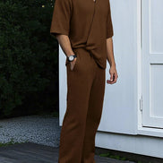 Man wearing a brown outfit standing on a wooden deck with a white wall and greenery in the background.