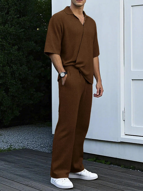 Man wearing a brown outfit standing on a wooden deck with a white wall and greenery in the background.