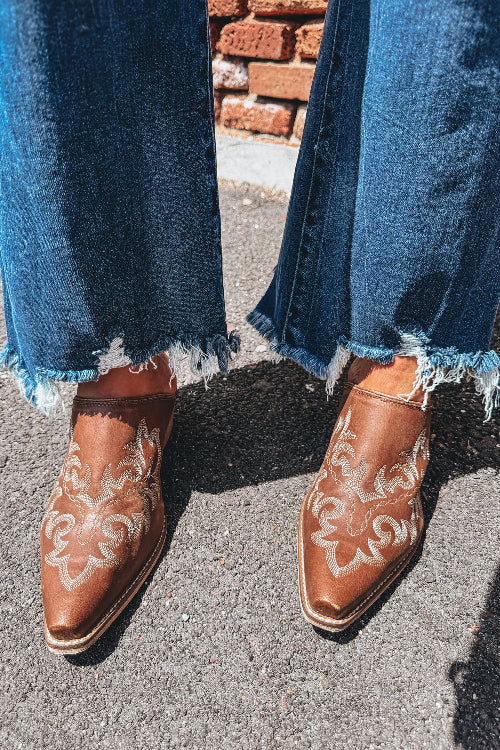 Brown cowboy boots with white embroidery worn with blue jeans on a pavement background