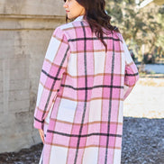 Woman wearing a pink plaid coat standing outdoors with trees and a building in the background.