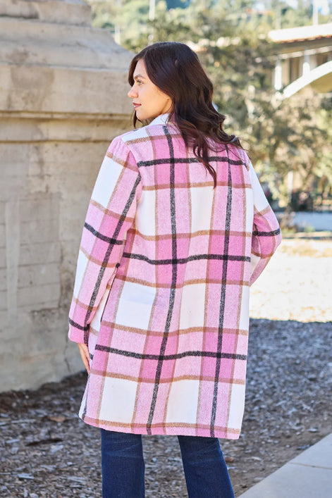 Woman wearing a pink plaid coat standing outdoors with trees and a building in the background.