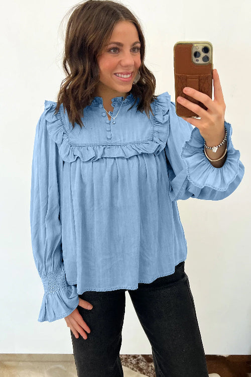 A woman taking a mirror selfie wearing a light blue long-sleeve blouse with ruffled details on the collar and cuffs, paired with black pants.