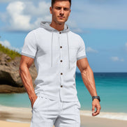 A man wearing a light gray short-sleeve button-front hoodie and matching jogger pants, standing on a sandy beach 
