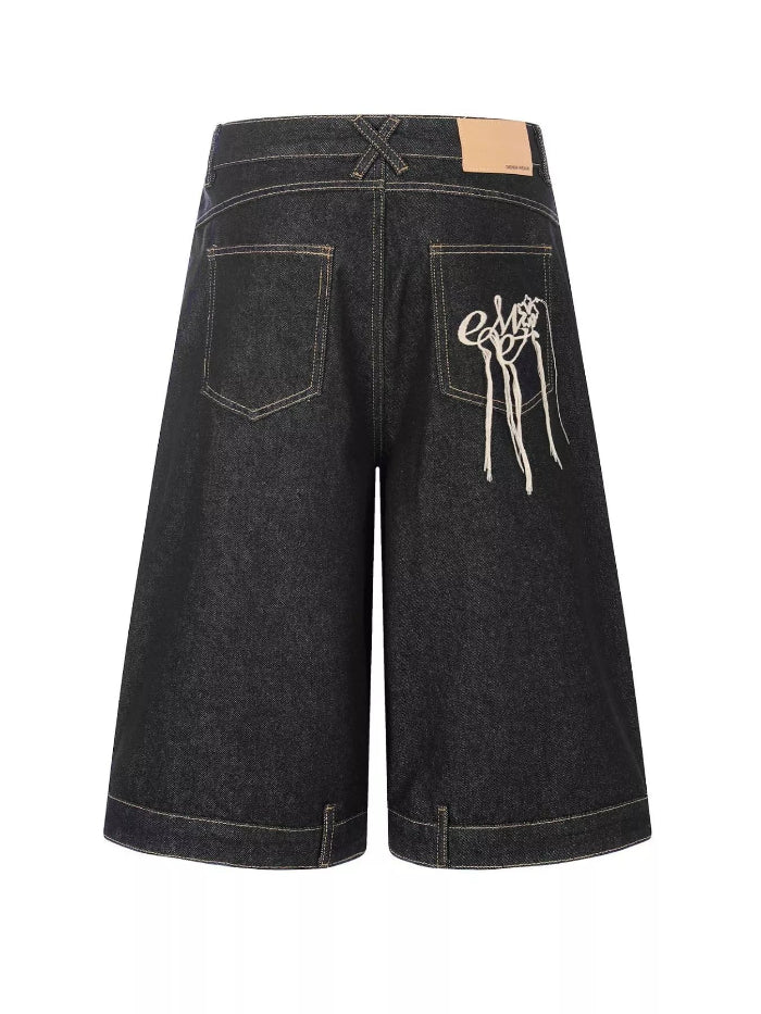 Back view of dark washed denim shorts featuring a white embroidered graphic on the right back pocket, a tan leather patch at the waistband, and contrast light tan stitching.