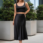 A woman wearing a black satin midi skirt with a flared hem, paired with a black crop top and clear high-heel sandals, 