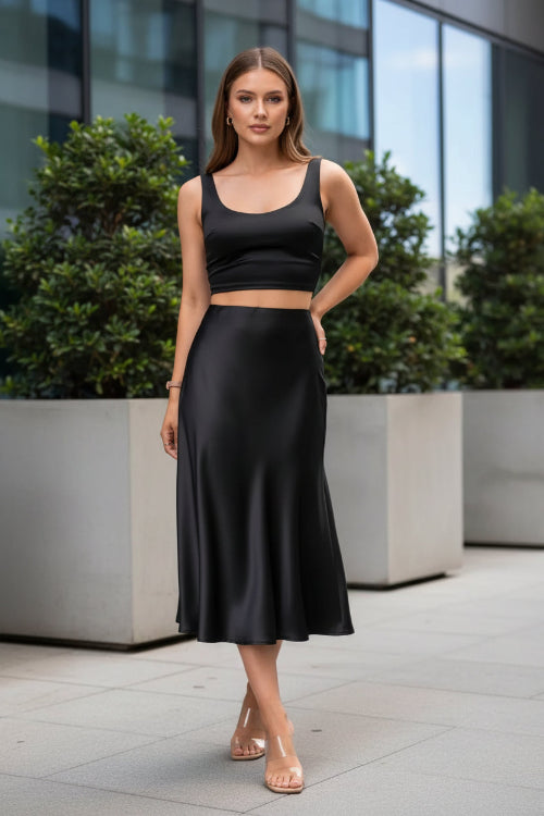 A woman wearing a black satin midi skirt with a flared hem, paired with a black crop top and clear high-heel sandals, 