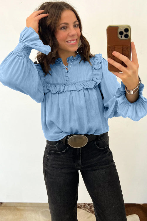 A woman taking a mirror selfie wearing a light blue long-sleeve blouse with ruffle details, paired with black jeans and a large silver belt buckle.