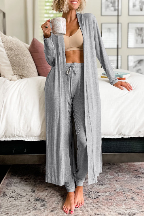 A woman stands in a bedroom wearing a matching gray lounge set consisting of a long open-front cardigan and drawstring sweatpants, 