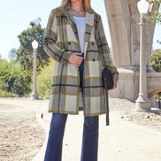 Woman wearing a plaid coat and blue jeans standing outdoors with architectural elements in the background.
