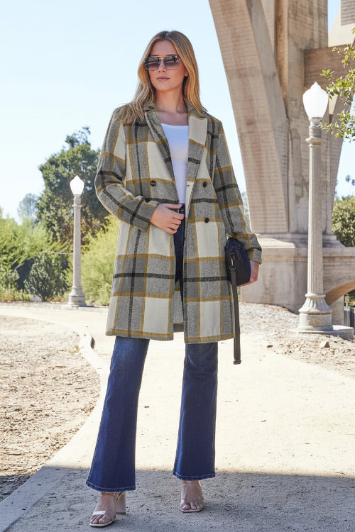 Woman wearing a plaid coat and blue jeans standing outdoors with architectural elements in the background.