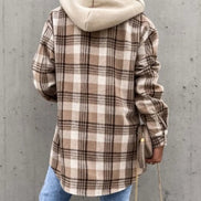 Person wearing a plaid shirt with a beige hoodie against a gray concrete wall
