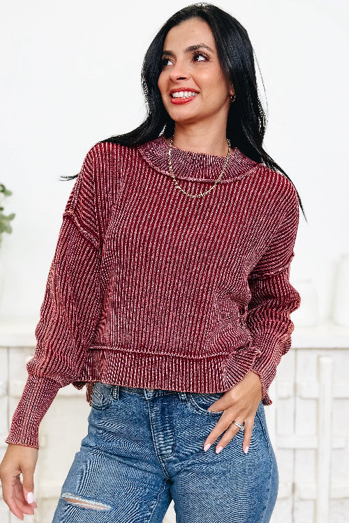 A woman wearing a ribbed long-sleeve sweater in a dusty rose or mauve color, paired with blue jeans, standing against a white brick wall.