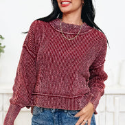A woman wearing a ribbed long-sleeve sweater in a dusty rose or mauve color, paired with blue jeans, standing against a white brick wall.