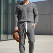 A man wearing a matching gray cable-knit sweater and jogger pants set, holding a brown wide-brim hat. 