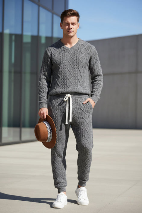 A man wearing a matching gray cable-knit sweater and jogger pants set, holding a brown wide-brim hat. 