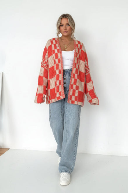 Person wearing a red and beige checkered cardigan over a white top and blue jeans on a plain background
