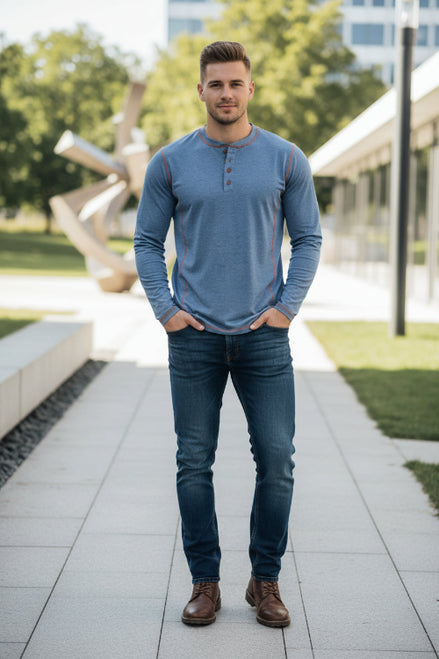 Man wearing a blue long-sleeve shirt and jeans standing on a sidewalk with greenery and buildings in the background.