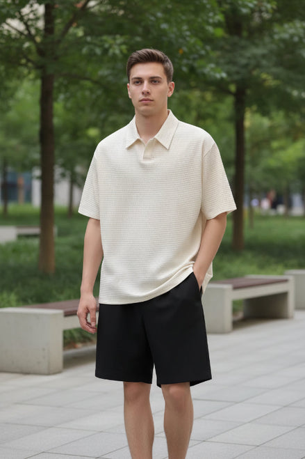 Person wearing a beige short-sleeve shirt and black shorts, holding a black bag against a gray wall.