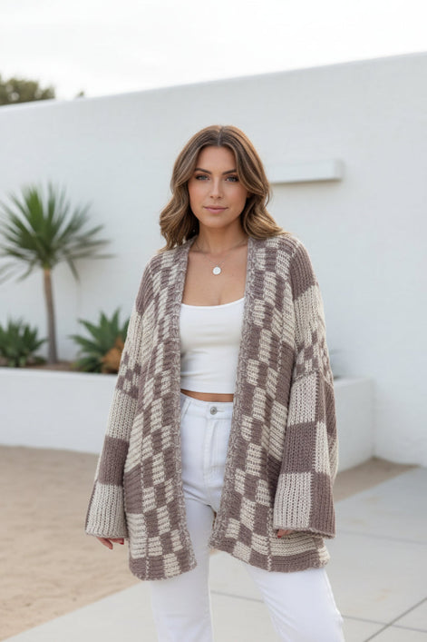 Woman wearing a checkered crochet cardigan over a white top and pants, standing indoors.