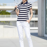 A woman wearing a navy and white horizontally striped short-sleeve polo shirt and a black baseball cap, holding a tennis racket against an outdoor background with barbed wire.