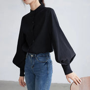 A woman wearing a black long-sleeve blouse with balloon sleeves and a buttoned mandarin collar, paired with blue jeans, standing against a light gray wall.