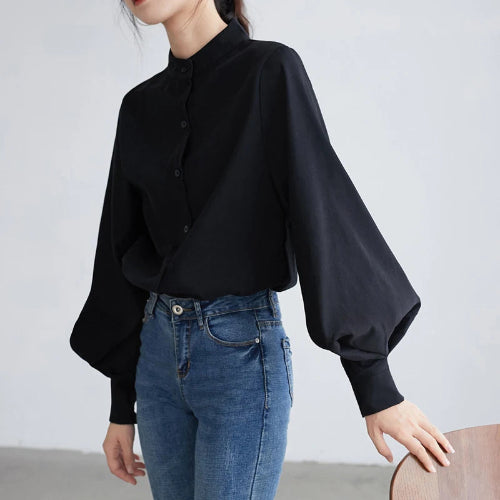 A woman wearing a black long-sleeve blouse with balloon sleeves and a buttoned mandarin collar, paired with blue jeans, standing against a light gray wall.