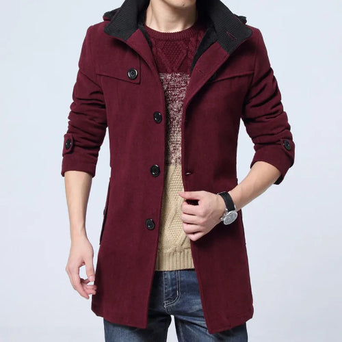 A man wearing a long burgundy wool-blend coat with a black collar, layered over a beige knit sweater and blue jeans, against a light gray studio background.