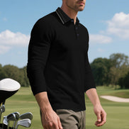 Angled front view of men's black long sleeve polo shirt with fine contrasting collar stripes and a button placket, modeled beside golf clubs on a golf course