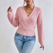 A woman wearing a light pink cable-knit wrap sweater with long sleeves and blue jeans, standing against a textured white wall.