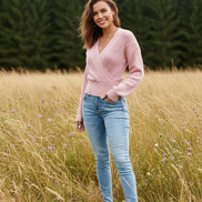 A woman wearing a light pink cable-knit wrap sweater with long sleeves and blue jeans, standing against a textured white wall.