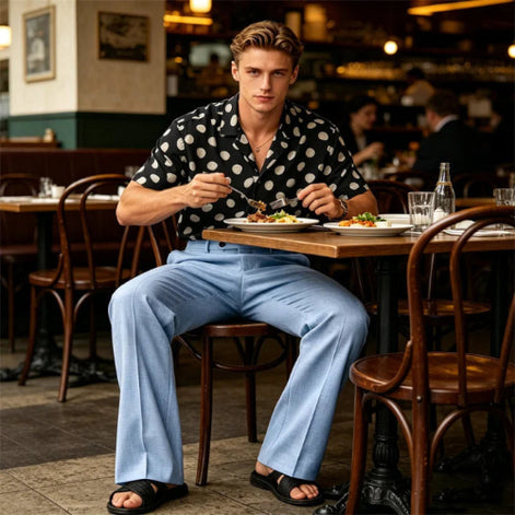 A man sits at a restaurant table wearing a black short-sleeve button-up shirt with white polka dots, light blue wide-leg trousers, and black slide sandals, holding a fork and knife over a plate of food.
