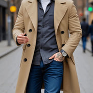 A man wearing a camel-colored double-breasted overcoat over a dark gray button-up shirt, blue jeans, and a white t-shirt, walking on a blurred city street.