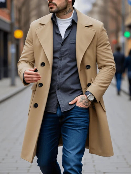 A man wearing a camel-colored double-breasted overcoat over a dark gray button-up shirt, blue jeans, and a white t-shirt, walking on a blurred city street.