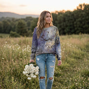 A woman wearing a long-sleeve crewneck sweatshirt with an abstract, painterly print in shades of purple, blue, gray, and gold, paired with blue jeans.