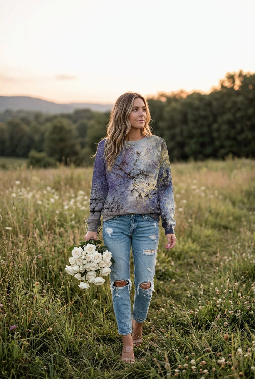 A woman wearing a long-sleeve crewneck sweatshirt with an abstract, painterly print in shades of purple, blue, gray, and gold, paired with blue jeans.