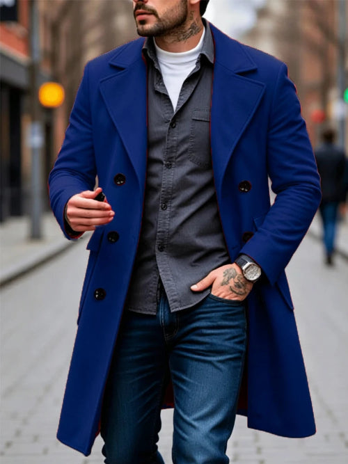 A man wearing a vibrant royal blue double-breasted overcoat over a dark gray button-down shirt, white t-shirt, and blue jeans, standing on a city street.
