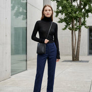 A person wearing a black sheer long-sleeve mock neck top and black wide-leg pants, carrying a small black crossbody bag, standing against a beige tiled wall.