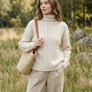 A woman wearing a cream-colored cable-knit sweater and beige pants, holding a black smartphone to take a mirror selfie. 