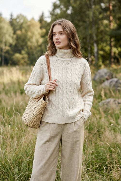 A woman wearing a cream-colored cable-knit sweater and beige pants, holding a black smartphone to take a mirror selfie. 
