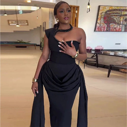 A woman modeling a black one-shoulder draped gown with beaded trim and a high-waisted silhouette in a modern indoor setting.