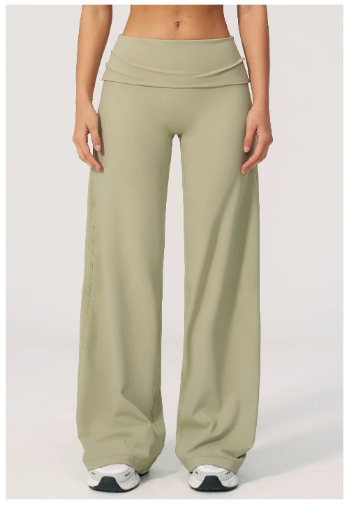 A model wearing high-waisted, wide-leg lounge pants in a muted sage green color, paired with white sneakers, shown from the waist down against a light gray studio background.