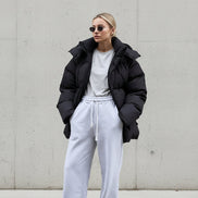 Person wearing a black puffer jacket and white pants standing against a gray wall.