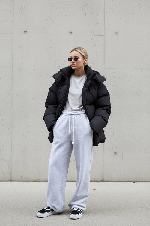 Person wearing a black puffer jacket and white pants standing against a gray wall.