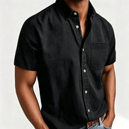 A man wearing a black short-sleeve button-up shirt with a chest pocket, paired with blue jeans and a brown leather belt, 